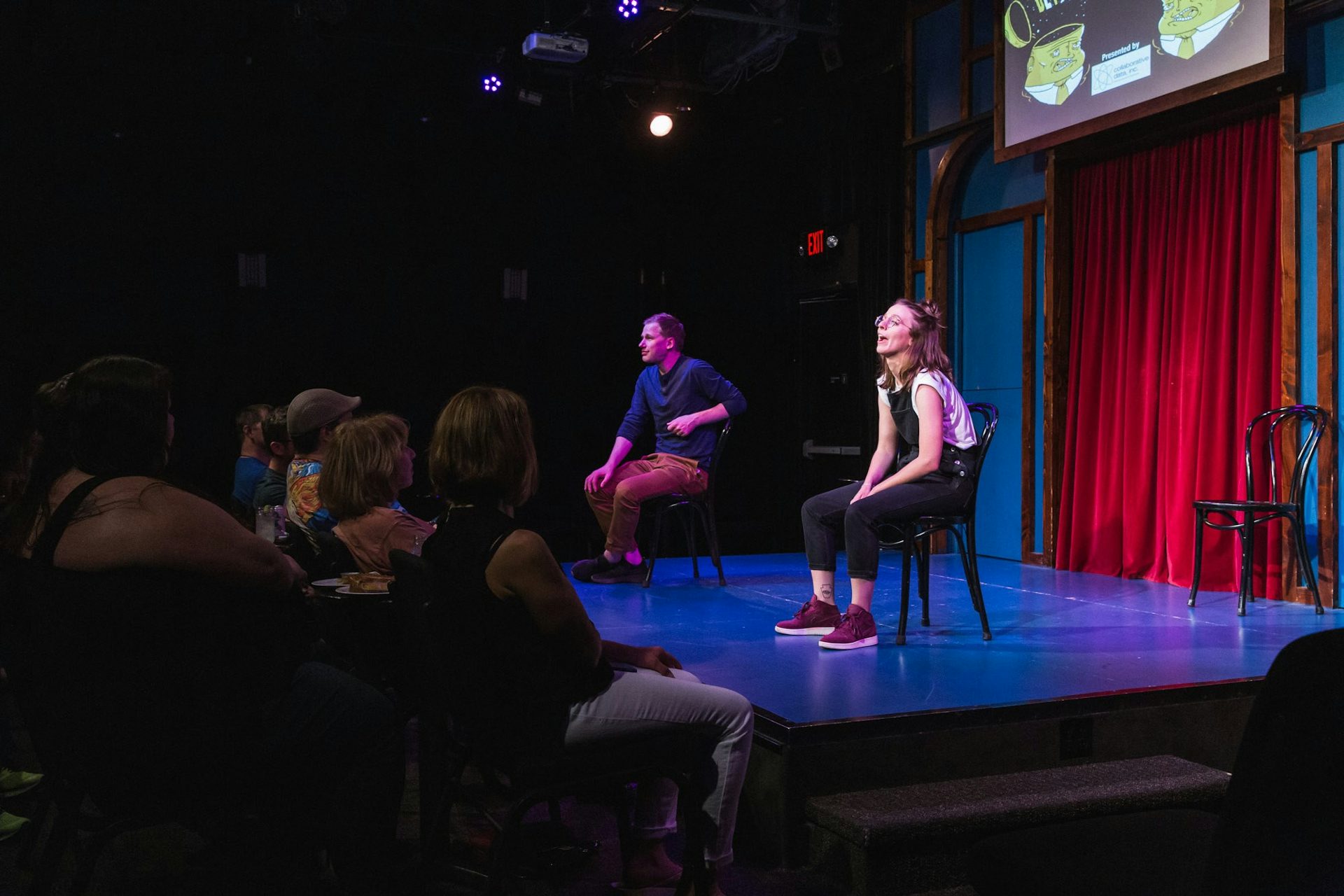 Jerome & Alex are on stage sitting on chairs in front of a crowd with a red curtain behind them. They and the chairs are pointed towards the crowd where Alex has a spotlight on them to deliver a soliloquy with Jerome off to her left.