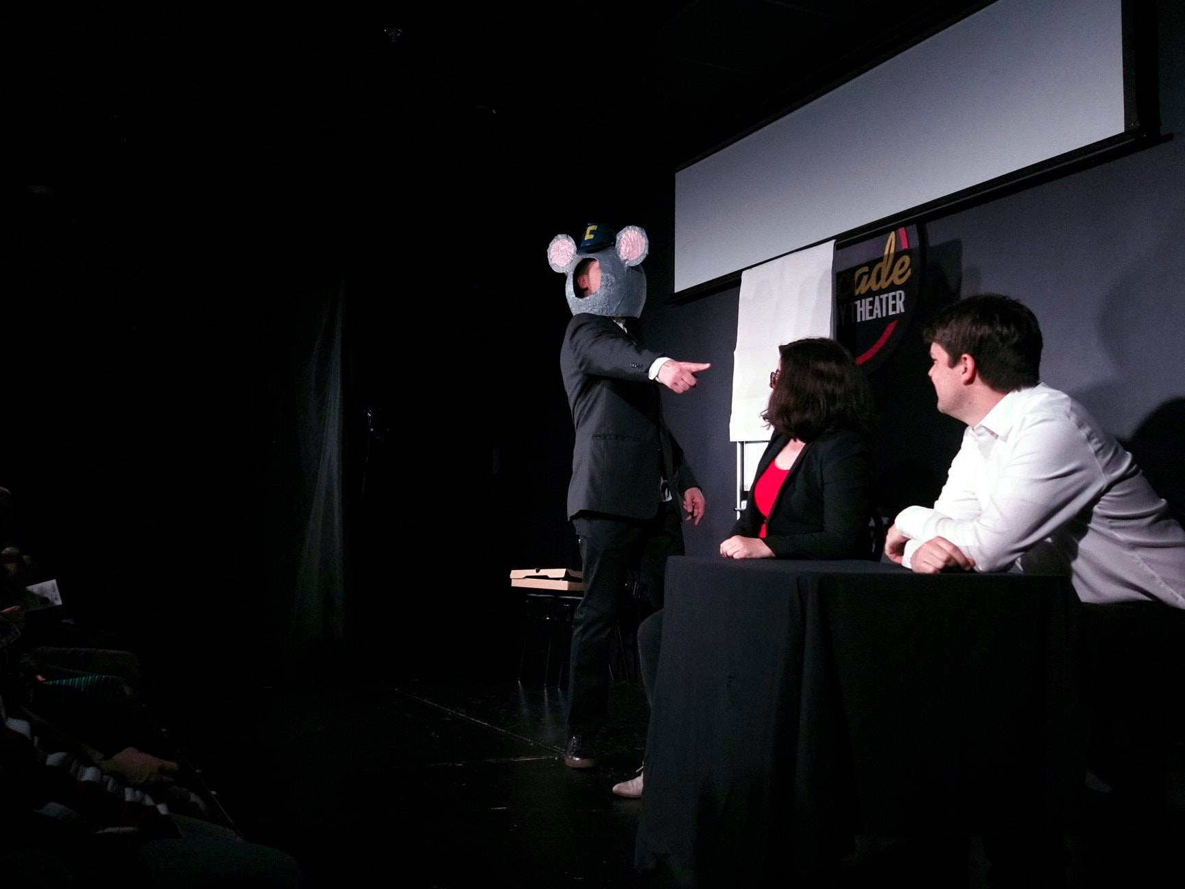 Jerome is wearing a black suit, with a paper mâché head of Charles Entertainment Cheese Junior. A blue duct-tap cap with a yellow “C” resides between two giant rat (mouse?) ears with a cut-out for his face. He is standing pointing an accusatory finger at two poor seated schlubs about to incur his wrath. Due to his stance and finger pointing you cannot see his face under the paper mâché rat head and just see his right ear and side cheek. There is an empty pizza box on a chair behind him.