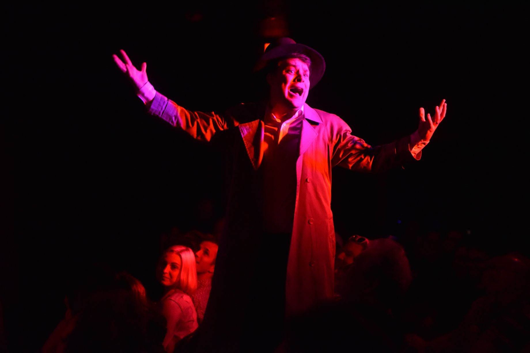 Jesse as Mr. Hyde washed in red light as he moves around the seated crowd. He is standing on a chair with his arms raised, mouth agape, with a top hat and trench coat on.